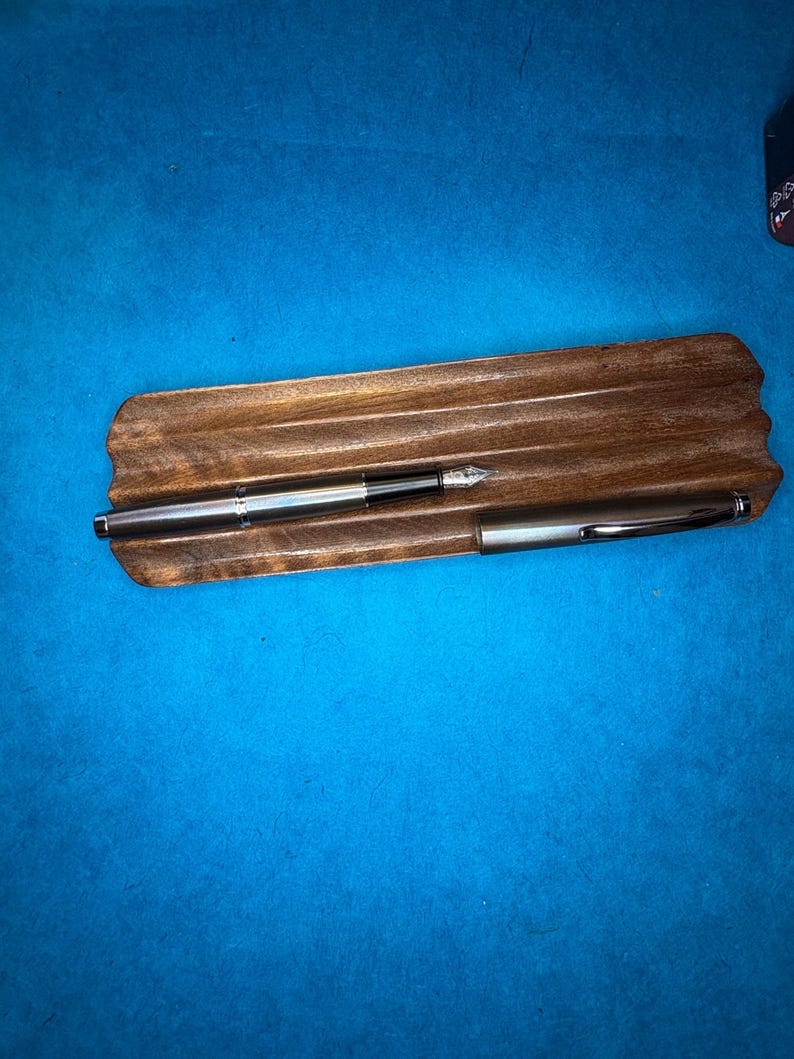 Puede incluir: Dos plumas estilogr&aacute;ficas plateadas descansan sobre un portal&aacute;pices de madera. Las plumas tienen un dise&ntilde;o elegante y moderno con detalles en negro. El soporte es de madera oscura con forma natural, sobre fondo azul.