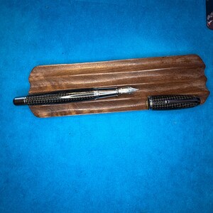 Puede incluir: Una pluma estilogr&aacute;fica con un cuerpo de fibra de carbono y detalles plateados descansa sobre una bandeja de madera para bol&iacute;grafos. La tapa de la pluma est&aacute; separada y colocada junto a la pluma. La bandeja es de color marr&oacute;n oscuro y el fondo es azul liso.