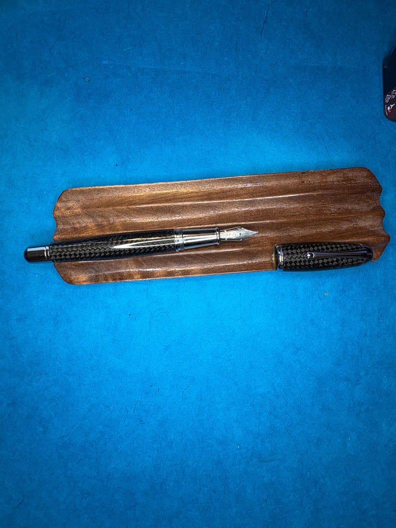 Puede incluir: Una pluma estilogr&aacute;fica con un cuerpo de fibra de carbono y detalles plateados descansa sobre una bandeja de madera para bol&iacute;grafos. La tapa de la pluma est&aacute; separada y colocada junto a la pluma. La bandeja es de color marr&oacute;n oscuro y el fondo es azul liso.