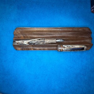 Puede incluir: Dos plumas estilogr&aacute;ficas descansan sobre una bandeja de madera. Las plumas tienen un patr&oacute;n veteado en tonos marrones y crema. La bandeja es de madera oscura con un dise&ntilde;o tallado. El fondo es azul liso.