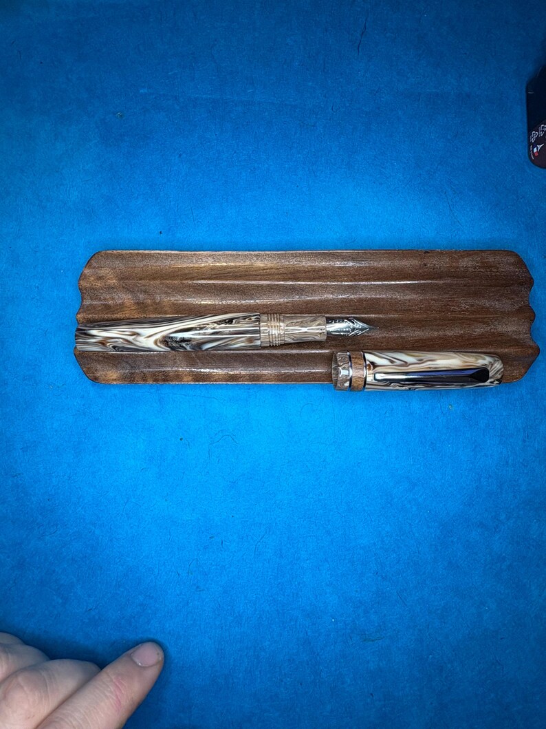 Puede incluir: Dos plumas estilogr&aacute;ficas descansan sobre una bandeja de madera. Las plumas tienen un patr&oacute;n veteado en tonos marrones y crema. La bandeja es de madera oscura con un dise&ntilde;o tallado. El fondo es azul liso.