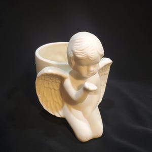 May include: White ceramic planter shaped like an angel with wings. The angel is kneeling and holding out its hand.