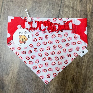 May include: A dog bandana in red and white, featuring a pattern of pink and red objects. It has a red polka dot section and a ruffled red trim. A tag with a dog logo and the text "Pip & Pup Co." is attached.
