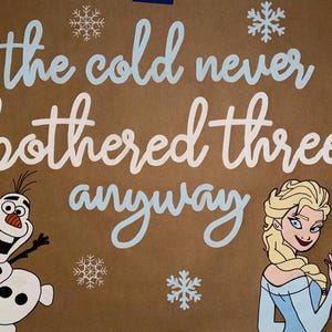 May include: A brown background with the phrase "the cold never bothered three anyway" in white and light blue script. The image features cartoon illustrations of Olaf the snowman and Elsa from the movie Frozen, along with snowflake accents.