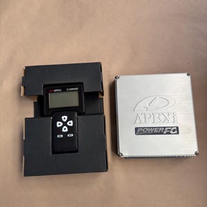 Puede incluir: Una unidad de control negra con una pequeña pantalla y botones, empaquetada en una caja negra, se encuentra junto a una caja metálica plateada con el logotipo APEXi Power FC. Los artículos están sobre una superficie de color canela.