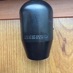 Op de afbeelding: Zwarte versnellingspookknop met het woord "NISMO" in reliëf. De knop heeft een afgeronde bovenkant en een taps toelopende basis. Het product is gemaakt van een solide, donker materiaal.