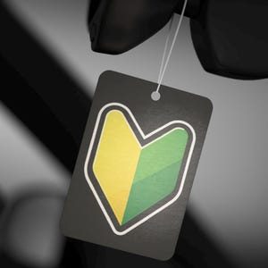 May include: A black tag with a white border, featuring a yellow and green beginner driver symbol. The tag is suspended by a white string, with a blurred black background.