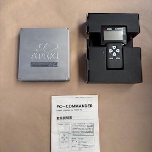 May include: A black and silver Apex Power FC Commander handheld console with a black and white screen and a five-button control pad. The console is in its original packaging, which is black with the Apex logo printed on it. The console is accompanied by a black and white instruction manual with the text "FC-COMMANDER HANDY CONSOLE for POWER FC 取扱説明書 APEXI" printed on it.