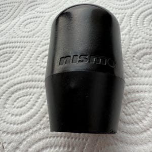 May include: Black shift knob with the embossed text "mismo". The knob has a rounded top and a tapered base. The surface appears to be matte. The shift knob is likely for a vehicle.