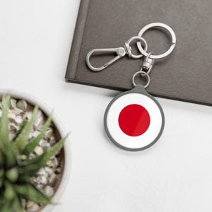 Puede incluir: Un llavero circular con fondo blanco y un círculo rojo sólido en el centro, que recuerda a la bandera nacional de Japón. El llavero incluye una anilla de metal plateado y un cierre de mosquetón unido a un marco de color gris oscuro.