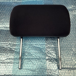 May include: A black car headrest with a rectangular cushion and two chrome support posts. The headrest is designed to provide comfort and support for the head and neck while driving.