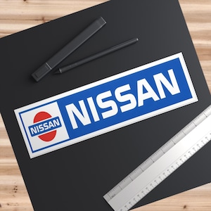 May include: A rectangular sticker with the word "NISSAN" in white, set against a blue background. The sticker also features the Nissan logo, a red circle with the word "NISSAN" in white. A ruler and two black pens are also visible.