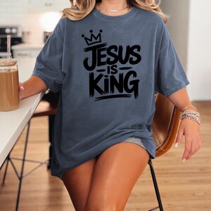 Pu&ograve; includere: Una maglietta blu denim con la scritta "JESUS IS KING" in nero, con una grafica a corona. La maglietta &egrave; di vestibilit&agrave; ampia e ha maniche corte. La parola "DENIM" &egrave; stampata in basso a sinistra nell'immagine.