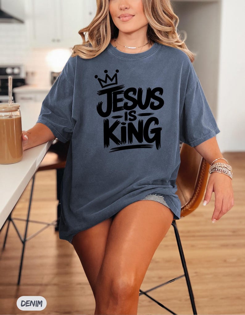 Pu&ograve; includere: Una maglietta blu denim con la scritta "JESUS IS KING" in nero, con una grafica a corona. La maglietta &egrave; di vestibilit&agrave; ampia e ha maniche corte. La parola "DENIM" &egrave; stampata in basso a sinistra nell'immagine.