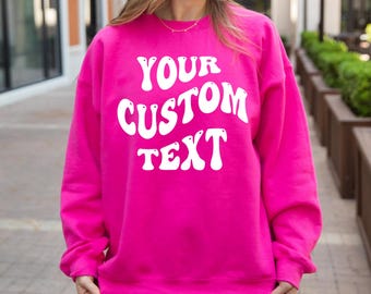 May include: A bright hot pink long-sleeve sweatshirt featuring the white text YOUR CUSTOM TEXT in a bold, wavy, retro-inspired font centered on the chest. The garment has a crew neckline, long sleeves with ribbed cuffs, and a ribbed hemline.