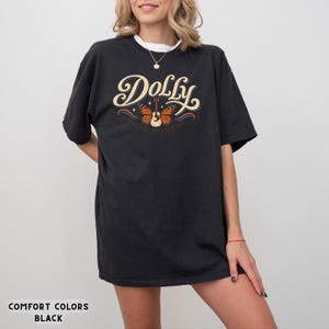 May include: A black short-sleeve t-shirt featuring the word Dolly in large cream-colored script lettering. Below the text is a graphic of an acoustic guitar with orange butterfly wings, accented by small stars and blue wavy lines.