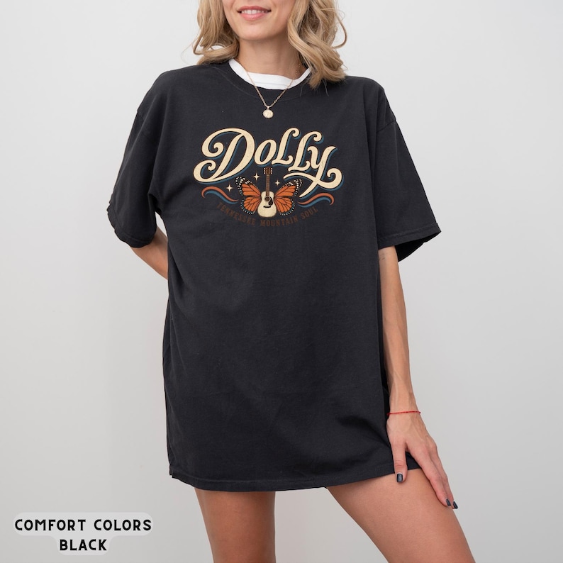 May include: A black short-sleeve t-shirt featuring the word Dolly in large cream-colored script lettering. Below the text is a graphic of an acoustic guitar with orange butterfly wings, accented by small stars and blue wavy lines.