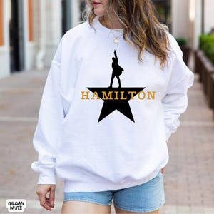 May include: White sweatshirt featuring a black silhouette of a person on a star, with the word "HAMILTON" in gold lettering. The sweatshirt is a Gildan White brand.