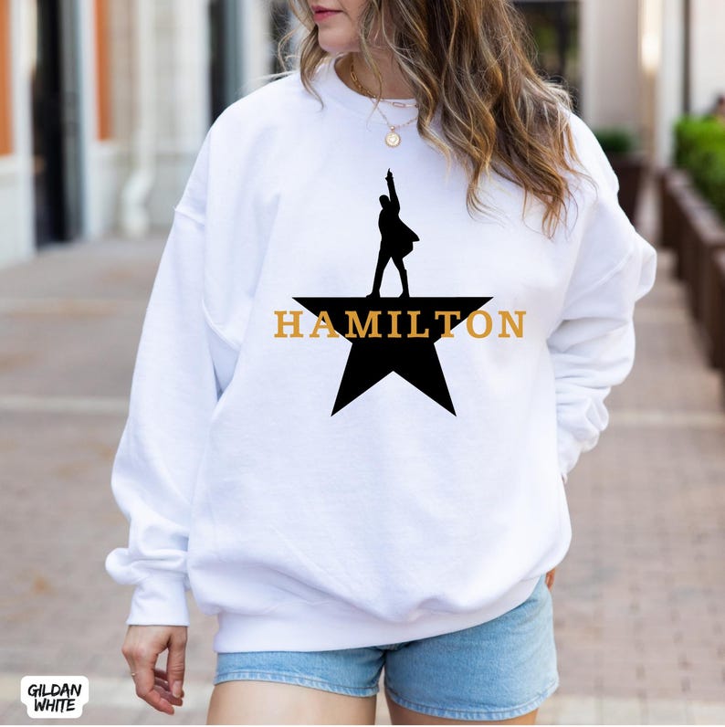 May include: White sweatshirt featuring a black silhouette of a person on a star, with the word "HAMILTON" in gold lettering. The sweatshirt is a Gildan White brand.