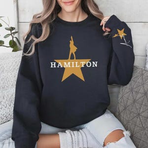 May include: A navy blue sweatshirt featuring a gold star with a silhouette of a person and the word "HAMILTON" in white. The right sleeve has a gold star and a signature in white.