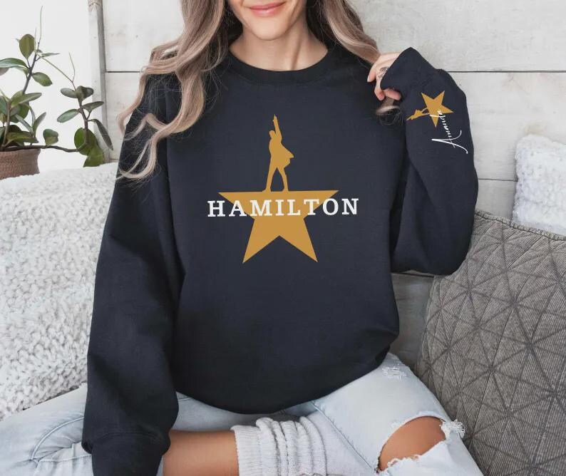 May include: A navy blue sweatshirt featuring a gold star with a silhouette of a person and the word "HAMILTON" in white. The right sleeve has a gold star and a signature in white.