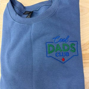 Puede incluir: Una sudadera azul de cuello redondo con el texto bordado "Cool Dads Club" en verde y azul, con una estrella roja. La sudadera est&aacute; doblada y sobre una superficie de madera clara.