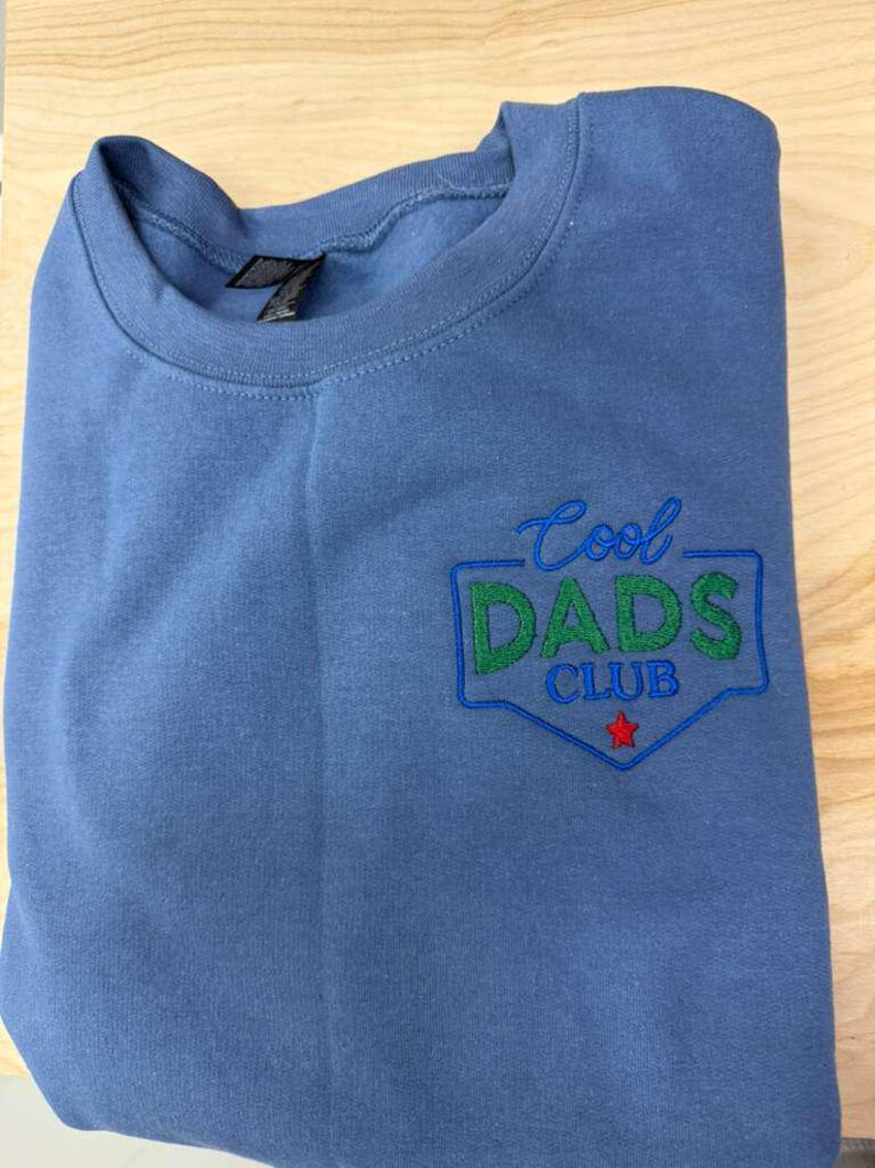 Puede incluir: Una sudadera azul de cuello redondo con el texto bordado "Cool Dads Club" en verde y azul, con una estrella roja. La sudadera est&aacute; doblada y sobre una superficie de madera clara.
