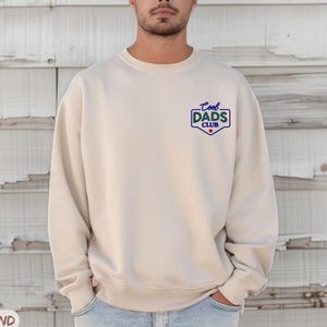 Puede incluir: Una sudadera de color arena con el texto "Cool Dads Club" en un emblema azul y blanco en el pecho. La sudadera es de color neutro y tiene cuello redondo.