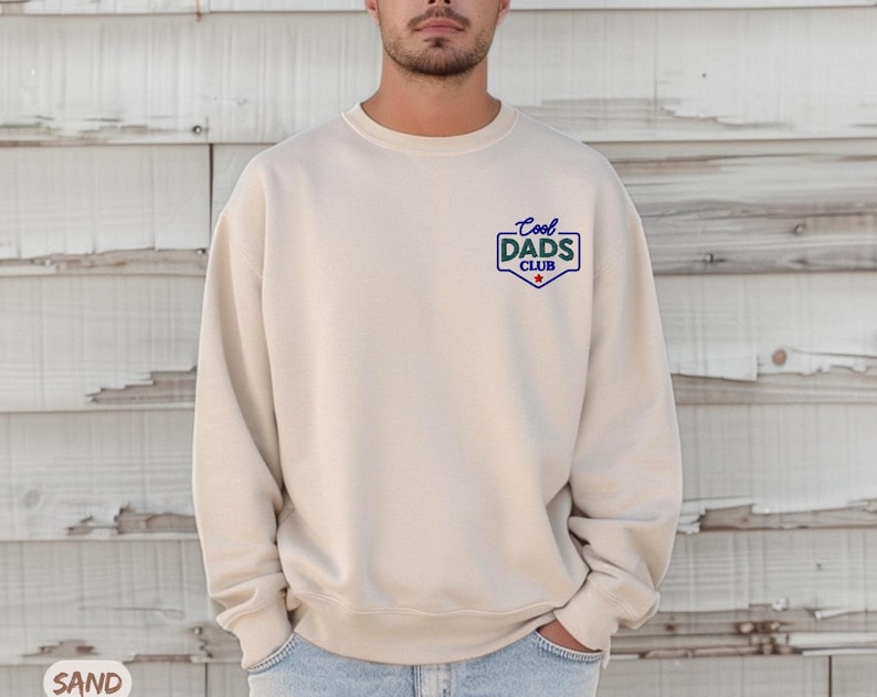 Puede incluir: Una sudadera de color arena con el texto "Cool Dads Club" en un emblema azul y blanco en el pecho. La sudadera es de color neutro y tiene cuello redondo.