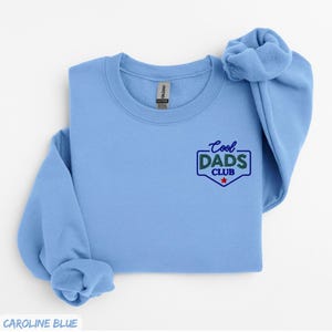 Puede incluir: Sudadera azul claro con el texto "Cool Dads Club" en un dise&ntilde;o verde y azul. La sudadera es de color liso y tiene cuello redondo. La manga est&aacute; doblada.
