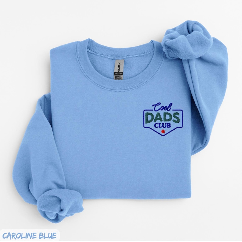 Puede incluir: Sudadera azul claro con el texto "Cool Dads Club" en un dise&ntilde;o verde y azul. La sudadera es de color liso y tiene cuello redondo. La manga est&aacute; doblada.