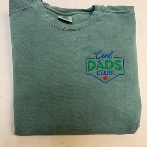 Puede incluir: Una sudadera verde salvia doblada con el texto bordado "Cool DADS CLUB" en verde y azul. Una peque&ntilde;a estrella roja est&aacute; debajo del texto. La sudadera est&aacute; hecha de un material suave.