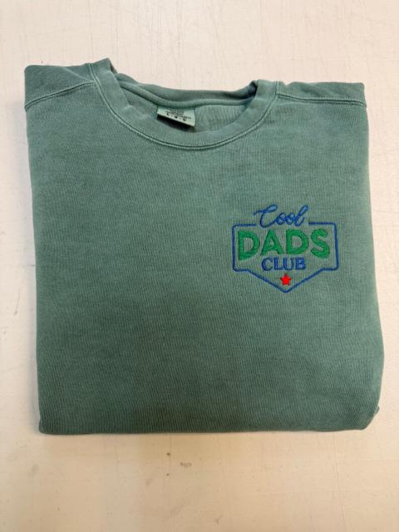 Puede incluir: Una sudadera verde salvia doblada con el texto bordado "Cool DADS CLUB" en verde y azul. Una peque&ntilde;a estrella roja est&aacute; debajo del texto. La sudadera est&aacute; hecha de un material suave.
