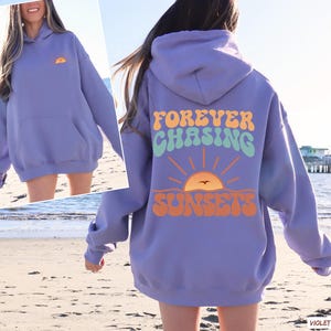 May include: A purple hoodie with the text "FOREVER CHASING SUNSETS" in a retro font with a sunset graphic. The hoodie has a large pocket on the front.