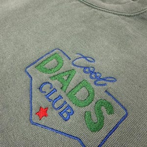 Puede incluir: Sudadera verde salvia con cuello redondo con un dise&ntilde;o bordado "Cool Dads Club". El dise&ntilde;o presenta letras verdes y azules, una estrella roja y un contorno azul.