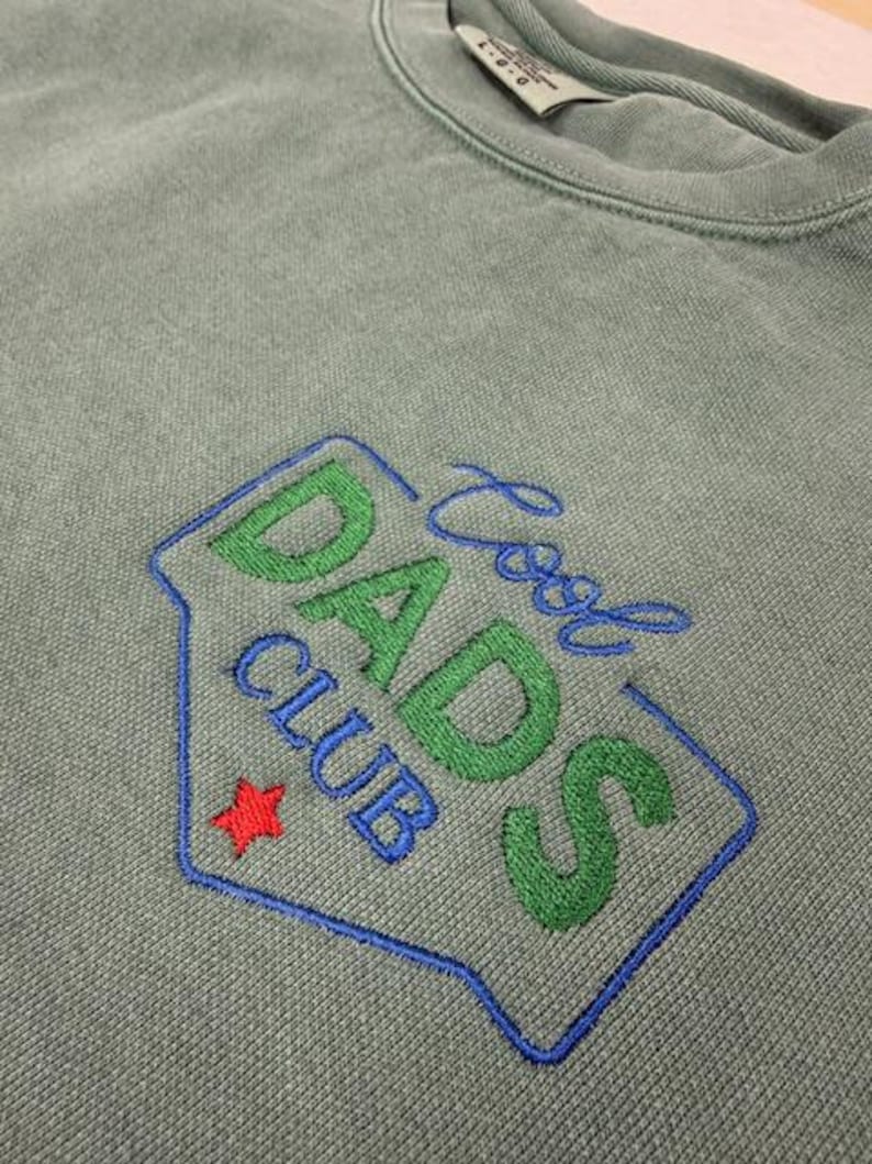 Puede incluir: Sudadera verde salvia con cuello redondo con un dise&ntilde;o bordado "Cool Dads Club". El dise&ntilde;o presenta letras verdes y azules, una estrella roja y un contorno azul.