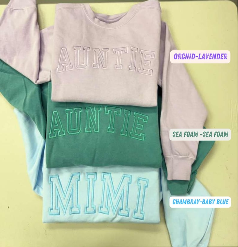 May include: Three sweatshirts in orchid-lavender, sea foam, and chambray-baby blue. The top two sweatshirts say "AUNTIE" in embroidered lettering. The bottom sweatshirt says "MIMI". The sweatshirts are folded and stacked.