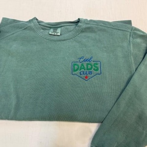 Puede incluir: Una sudadera verde salvia con cuello redondo y el texto bordado "Cool Dads Club" en azul y verde. El dise&ntilde;o tiene forma de escudo con una estrella roja en la parte inferior. La sudadera tiene mangas largas y un corte holgado.