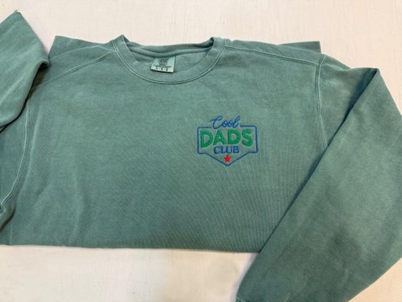 Puede incluir: Una sudadera verde salvia con cuello redondo y el texto bordado "Cool Dads Club" en azul y verde. El dise&ntilde;o tiene forma de escudo con una estrella roja en la parte inferior. La sudadera tiene mangas largas y un corte holgado.