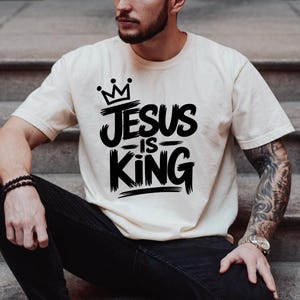 Pu&ograve; includere: T-shirt color crema con la scritta "JESUS IS KING" in grassetto nero, sormontata da una corona. La maglietta a maniche corte sembra essere realizzata in un materiale morbido.
