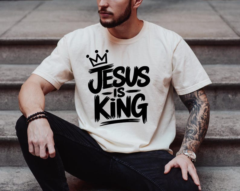 Pu&ograve; includere: T-shirt color crema con la scritta "JESUS IS KING" in grassetto nero, sormontata da una corona. La maglietta a maniche corte sembra essere realizzata in un materiale morbido.
