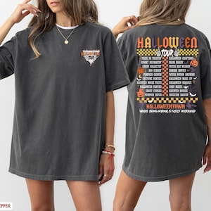 May include: Dark gray Halloween Tour t-shirt. The front features a triangular graphic with the words "Halloween Tour". The back of the shirt lists Halloween-themed activities in orange, yellow, and purple. The text "Halloweentown" is also printed on the back.