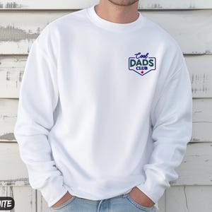 Puede incluir: Sudadera blanca con el texto "Cool Dads Club" en azul y verde en el pecho. La sudadera tiene cuello redondo y mangas largas. La palabra "WHITE" est&aacute; impresa en un recuadro blanco en la parte inferior izquierda.
