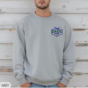 Puede incluir: Sudadera gris deportiva con el texto "Cool Dads Club" en azul y verde en el pecho. El texto est&aacute; dentro de un escudo con una estrella roja. Cuello redondo y mangas largas.