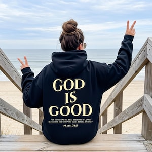 May include: Black hoodie with the words "GOD IS GOOD" in a gold color on the back. Below the text is a Bible verse from Psalm 34:8. The person is sitting on wooden steps near the beach.
