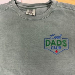 Puede incluir: Una sudadera verde salvia con cuello redondo y la frase bordada "Cool DADS Club" en azul y verde. El dise&ntilde;o incluye una estrella roja. La sudadera tiene una etiqueta que dice L-G.