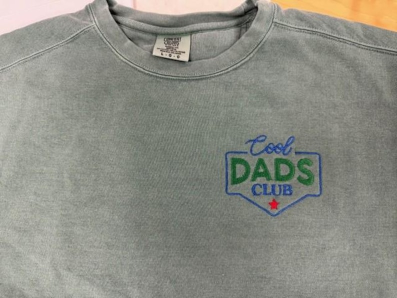 Puede incluir: Una sudadera verde salvia con cuello redondo y la frase bordada "Cool DADS Club" en azul y verde. El dise&ntilde;o incluye una estrella roja. La sudadera tiene una etiqueta que dice L-G.