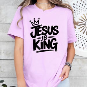 Pu&ograve; includere: T-shirt lilla con la scritta "JESUS IS KING" in nero, con un font a pennello. Il design include una corona sopra la parola "Jesus". La maglietta &egrave; a maniche corte.