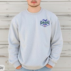 Puede incluir: Sudadera gris claro con el texto "Cool Dads Club" en un dise&ntilde;o azul y verde. La sudadera tiene cuello redondo y mangas largas. El texto est&aacute; en un dise&ntilde;o tipo insignia con una estrella roja.