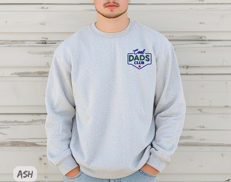 Puede incluir: Sudadera gris claro con el texto "Cool Dads Club" en un dise&ntilde;o azul y verde. La sudadera tiene cuello redondo y mangas largas. El texto est&aacute; en un dise&ntilde;o tipo insignia con una estrella roja.